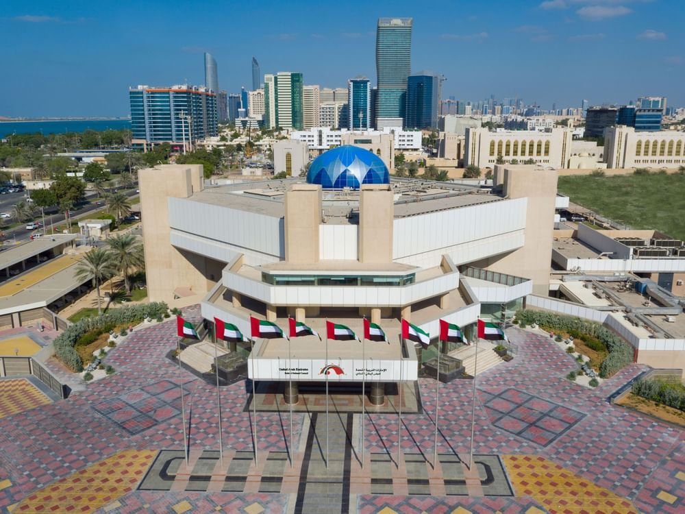An aerial view of a UAE government building with national flags on the forecourt, the Abu Dhabi skyline beyond