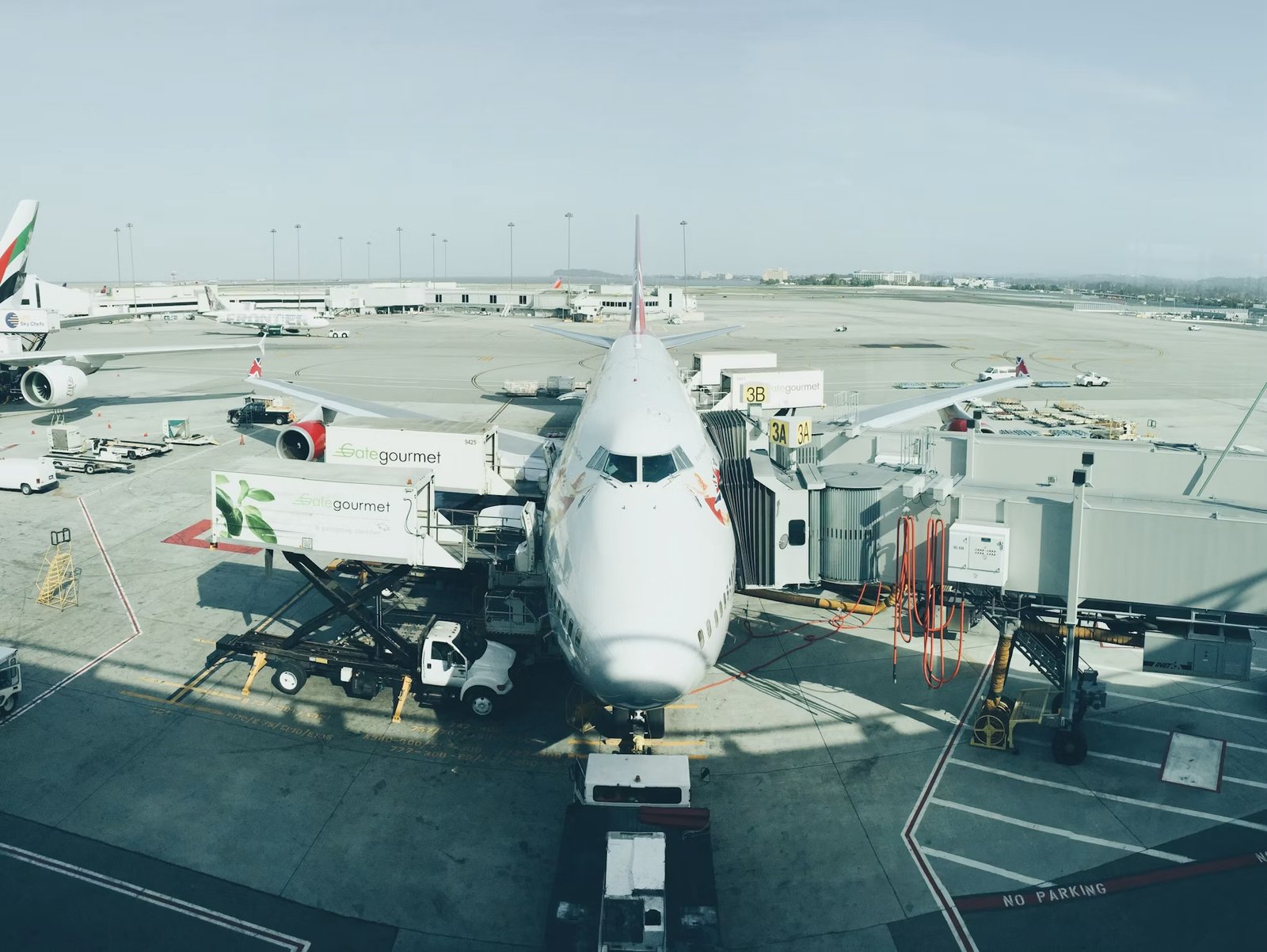 A widebody airliner parked at an airport gate, jet bridge and catering vehicles alongside